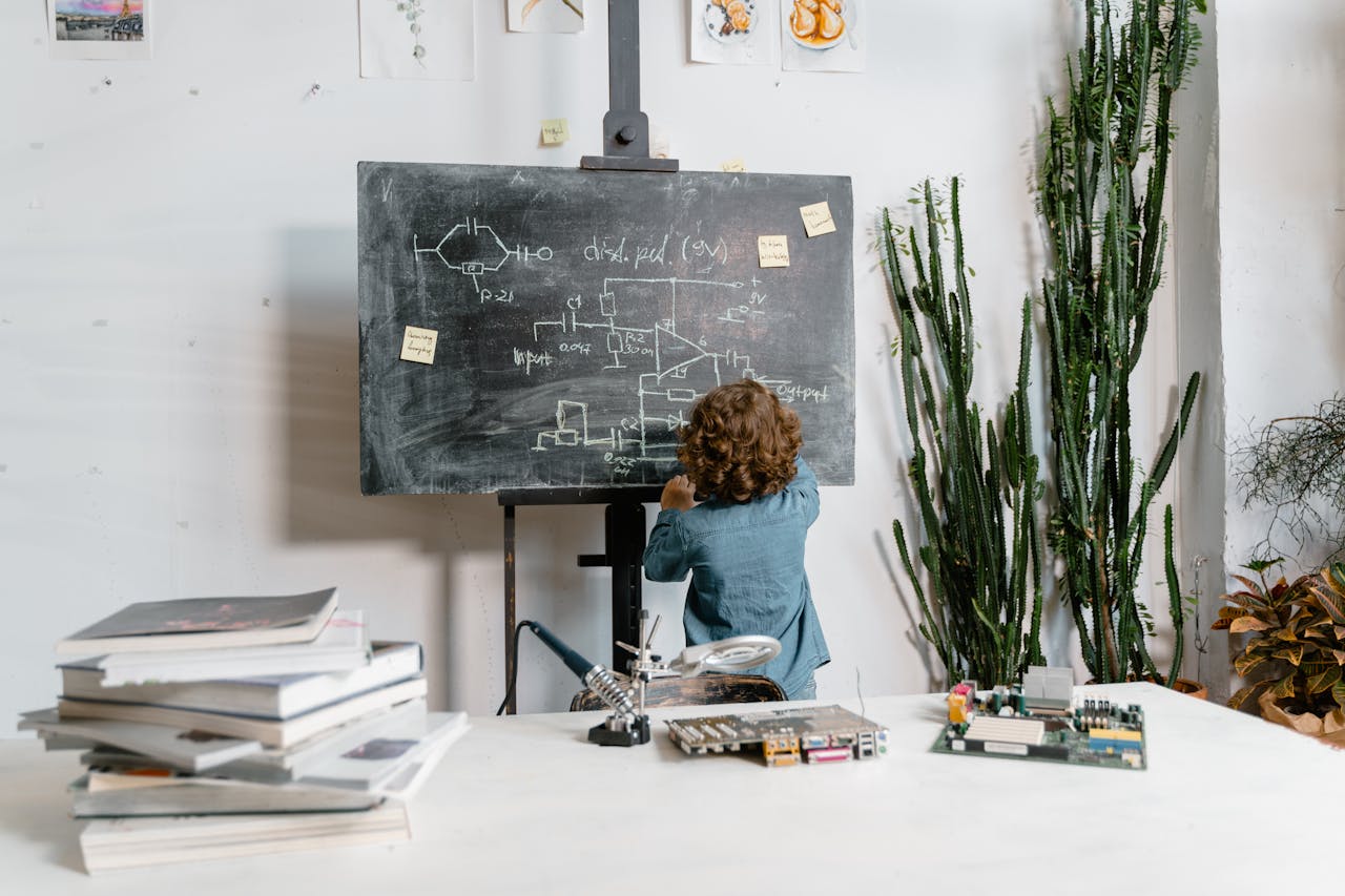 Curious young boy drawing STEM concepts on a blackboard indoors, surrounded by educational materials.